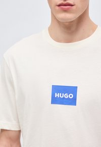 Κοντινή λήψη ενός ατόμου που φοράει λευκό μπλουζάκι με μπλε τετράγωνο και λευκά γράμματα "HUGO" στο στήθος.