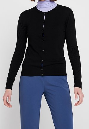 Femme portant un cardigan noir cintré à boutons par-dessus un col roulé lavande et un pantalon bleu ample, debout, les bras légèrement écartés.