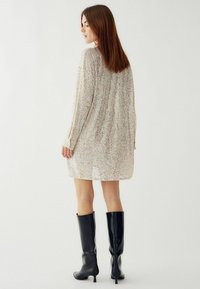 Blizganti kreminė ilgomis rankovėmis su laisvu siluetu ir blizgučių tekstūra, derinama su juodais iki kelių aukštakulniais.