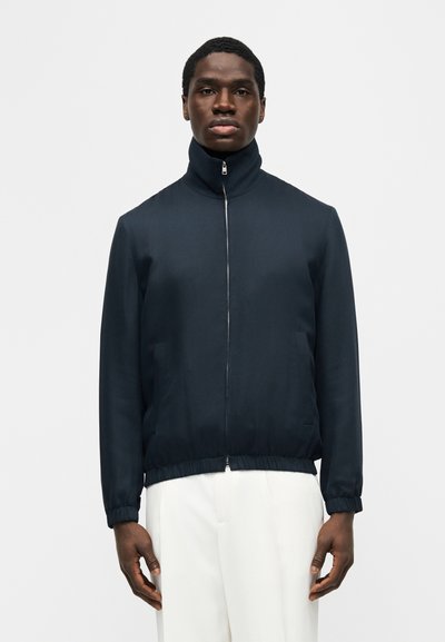 Homme portant une veste zippée bleu marine avec un col montant et des poignets élastiques, associé à un pantalon blanc, debout devant un fond uni.