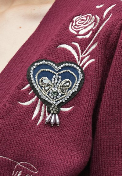 Broszka w kształcie serca z niebieskiej tkaniny, ozdobiona srebrnymi i czarnymi elementami oraz perłopodobnymi zawieszkami. Noszona na bordowym swetrze z białą kwiatową wyszywanką.