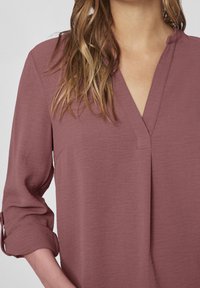 Blouse rose avec col en V, fabriquée en matière légère. Manches trois-quarts avec un design retroussé. Texture lisse.