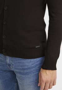 Mørkebrun strikket cardigan med knappelukking foran, ribbestrikkede mansjetter og kant, og et lite logomerke nederst til venstre. Båret over lyseblå jeans.