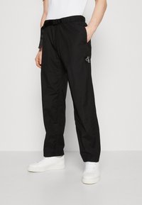 Černé cargo kalhoty s pružným pasem na šňůrku, bočními kapsami a nášivkou s logem. Vyrobené z lehkého materiálu. Nosené s bílými teniskami.