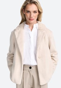Veste en fausse fourrure beige clair avec un col textur�é et bouclé, fermeture à un bouton et coupe décontractée, associée à une chemise blanche et un pantalon beige.