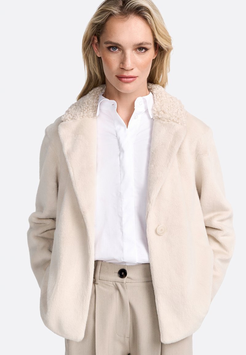 Veste en fausse fourrure beige clair avec un col texturé et bouclé, fermeture à un bouton et coupe décontractée, associée à une chemise blanche et un pantalon beige.