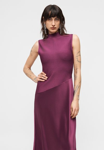 Γυναίκα με σκούρα μαλλιά μήκους ως τους ώμους, φορώντας μακρύ μωβ φόρεμα χωρίς μανίκια, κρυστάλλινα σκουλαρίκια-σταγόνες και εμφανίζοντας πολλά τατουάζ στα χέρια.