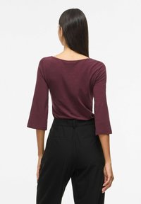 Tumši sarkans tops ar apaļu kaklu un 3/4 piedurknēm, izgatavots no mīksta auduma. Saskaņots ar melnām augstās jostasvietas biksēm ar sānu kabatām.