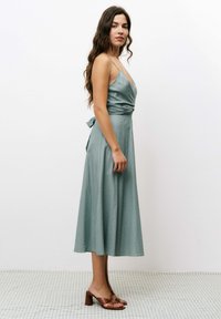 Une robe portefeuille en bleu sarcelle clair avec un décolleté en V profond, des bretelles fines et un lien à la taille. Elle présente un ourlet midi fluide et une texture subtile.