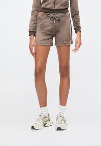 Γυναίκα που φοράει καφέ βελουτέ σορτς και αντίστοιχο τοπ, λευκές κάλτσες και μπεζ αθλητικά παπούτσια Nike, στέκεται μπροστά σε λευκό φόντο.