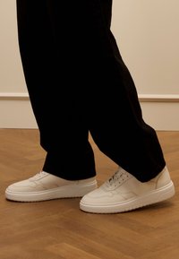 Weiße Ledersneaker mit glatter Textur, runder Zehenform und flacher Gummisohle, kombiniert mit schwarzen, locker sitzenden Hosen auf einem Holzboden.