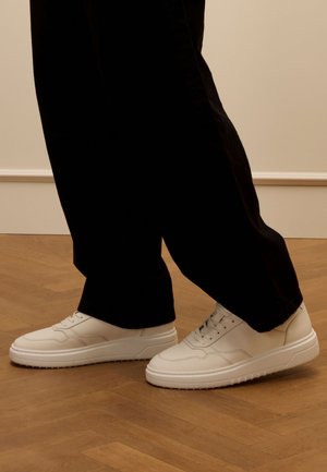 White leather sneakers with a smooth texture, round toe, and flat rubber sole, paired with black, loose-fitting trousers on a wooden floor.