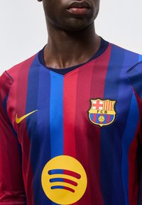 Άνδρας που φοράει φανέλα ποδοσφαίρου της FC Barcelona με κόκκινες και μπλε ρίγες, κίτρινο λογότυπο Nike και κίτρινο λογότυπο χορηγού Spotify.