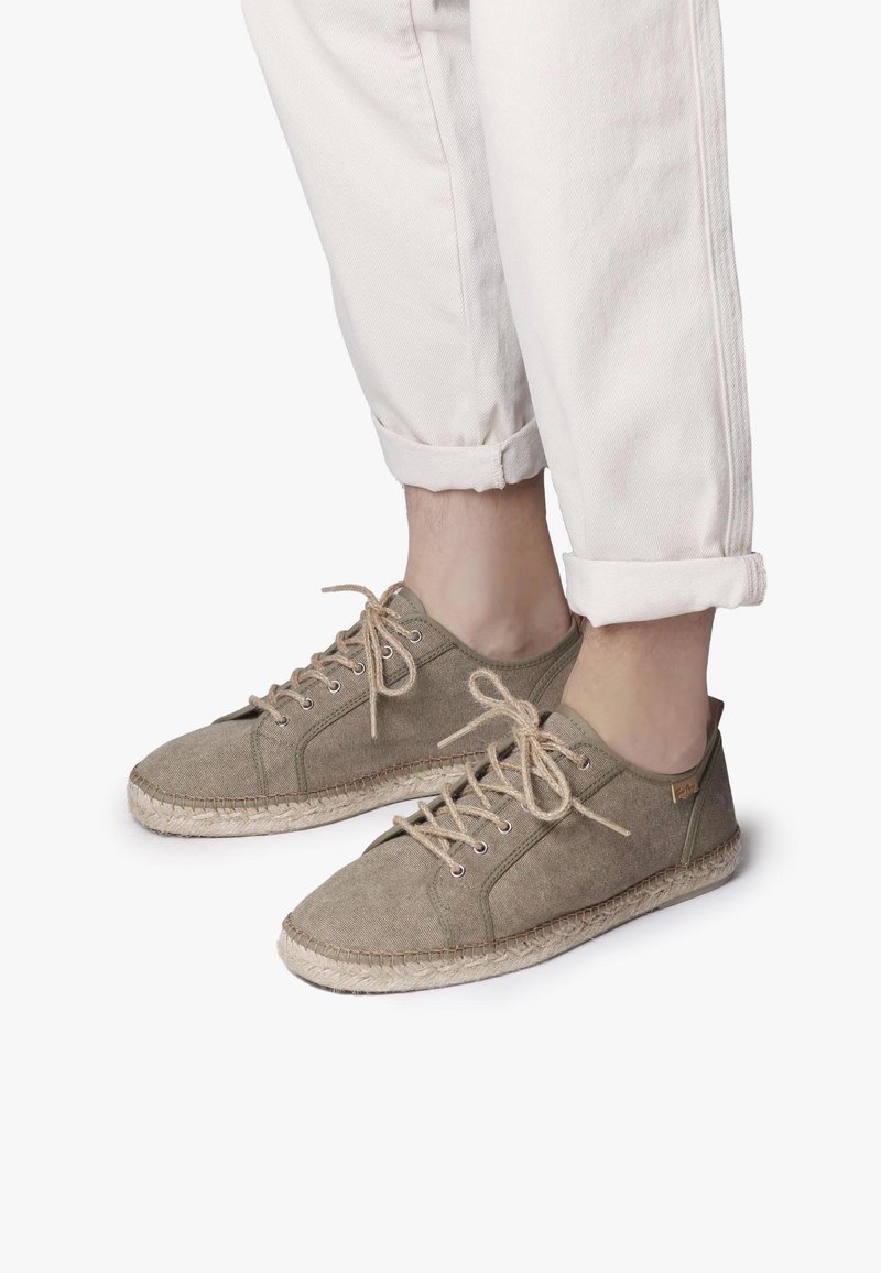 Personne portant un pantalon blanc retroussé et des chaussures en toile beige à lacets avec semelles en corde tressée sur fond blanc.