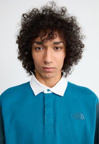 Türkiz póló fehér gallérral, gombos záródással és hímzett "The North Face" logóval a bal mellrészen. Göndör haj és septum piercing.