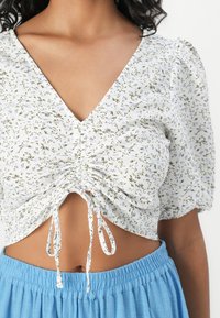 Vit blommig crop top med djup V-ringning, samlad framtill och knytningar; har korta puffärmar och en ljusblå, elastisk midjetröja.