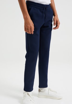 Personne portant un pantalon de jogging texturé bleu marine, des chaussettes blanches et des baskets blanches, debout devant un fond clair uni.