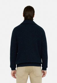 Granatowy sweter teksturowany z kolorowymi plamkami, z kołnierzem szalowym i prążkowanymi mankietami. Noszony na jasnych spodniach. Widok z tyłu.
