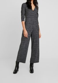 Combinaison en tricot grise avec un décolleté en V et des manches trois-quarts, présentant un pantalon large et un tissu texturé. Des bottines noires ajoutent la touche finale.