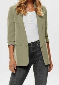 Olive green blazer with structured lapels, two front pockets, and rolled sleeves, layered over a white top and black jeans.