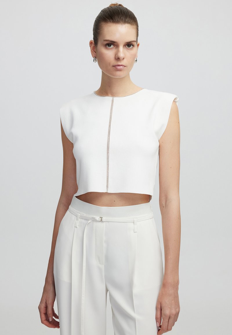 Witte cropped top met kapmouwen, featuring een centrale decoratieve naad. Gecombineerd met hoge witte broeken, gemaakt van een gladde stof, en een riem.