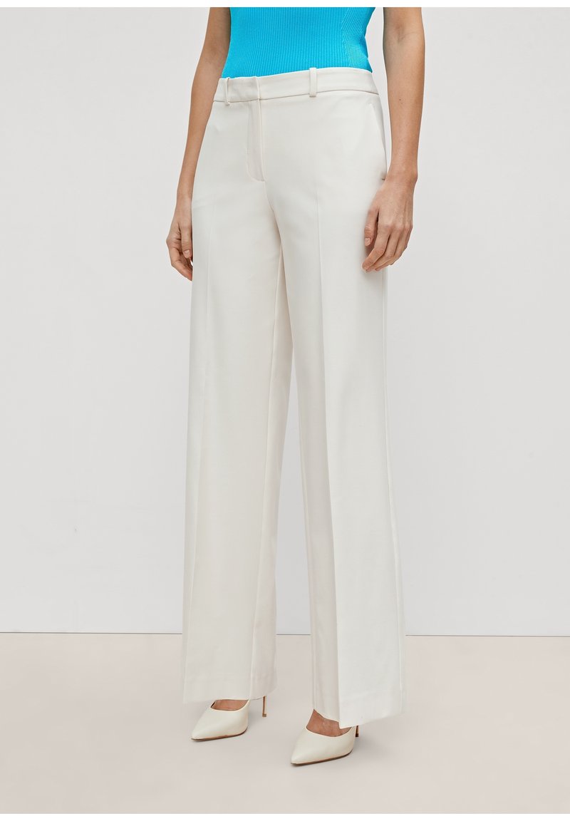 Femme portant un pantalon blanc taille haute à jambes larges, associé à des talons blancs pointus et un haut sans manches bleu vif rentré.