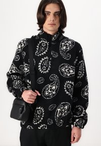 Svart og hvit mønstrete fleecegenser med høy krage, lange ermer og frontglidelås. Stylet med en liten svart crossover-bag.