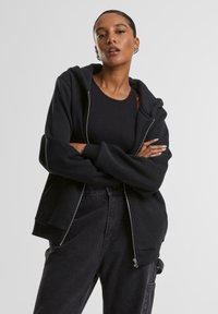 Nő keresztbe tett karokkal, fekete trikóban, túlméretezett cipzáras kapucnis pulóverben és magas derekú sötét farmerral, egyszínű háttér előtt.