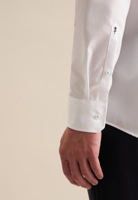 Manica di camicia bianca con polsino a bottone e ricamo di una piccola rosa nera sul lato esterno. Tessuto liscio, vestibilità sagomata.