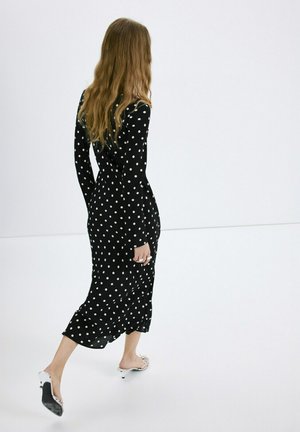 Mujer de cabello largo caminando de espaldas, vistiendo un vestido midi negro con lunares blancos y sandalias de tacón blancas sobre un suelo blanco.