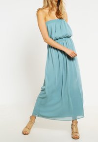 Femme portant une robe maxi sans manches, bleu clair, avec une taille élastique, accompagnée de sandales à talons hauts ouvertes beiges, se tenant devant un fond blanc.