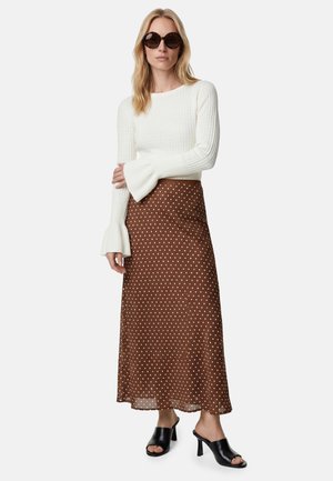 Kobieta nosząca ciemne okulary przeciwsłoneczne, biały prążkowany top z długim rękawem i rozszerzanymi mankietami, brązową spódnicę midi w kropki oraz czarne sandały na obcasie typu mule.