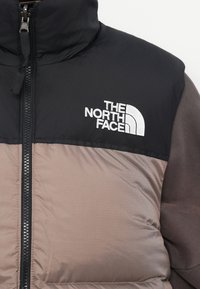 Puffjakas ar melnu augšējo daļu un brūnu apakšējo daļu, ar sudraba rāvējslēdzēju un balto The North Face logotipu uz krūtīm.