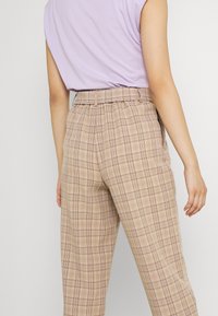 Person som bär beige rutiga högmidjade byxor och en instoppad ljuslila ärmlös topp, visad bakifrån.