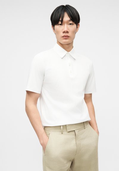 Νεαρός με μαύρα μαλλιά, ντυμένος με λευκό κοντομάνικο πόλο και μπεζ παντελόνι, στέκεται με τα χέρια στις τσέπες μπροστά σε απλό φόντο.