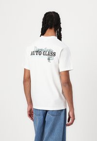 White cotton t-shirt with a graphic print on the back reading "Downtown AUTO GLASS" in black and teal. Short sleeves. Casual fit.