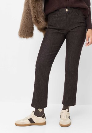 Persona que lleva pantalones oscuros de corte recto, zapatillas beige y marrones, calcetines oscuros con patrones de corazones y sostiene un abrigo de piel marrón colgado sobre un brazo.
