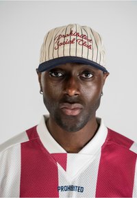 Baseball cap med cremefarvet stof, lodrette marineblå striber og rød broderet tekst. Bæres med en rød og hvid mønstret trøje med krave.