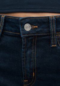 Κοντινό πλάνο σκούρων μπλε τζιν με πορτοκαλί ραφές και μεταλλικό κουμπί στη μέση με την επιγραφή "GAP DENIM EST. 1969".