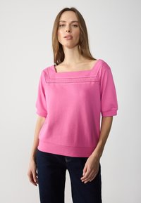 Top in magenta a maglia con scollatura quadrata, maniche corte e dettagli decorativi a occhio lungo lo scollo. Indossato con jeans scuri e larghi.