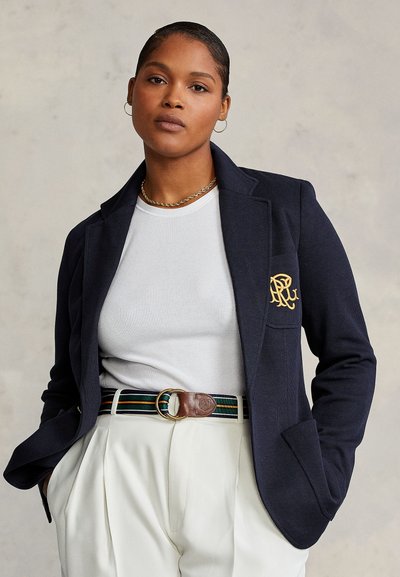 Jeune femme aux cheveux tirés en arrière, portant un blazer bleu marine, un t-shirt blanc côtelé, une ceinture rayée, un pantalon blanc taille haute et des boucles d’oreilles créoles dorées.