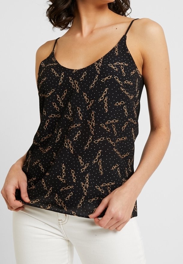 Femme portant un top noir sans manches avec des fines bretelles et un motif chaîne et points dorés, associé à un pantalon blanc.