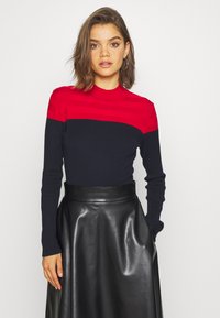 Un pull côtelé rouge et marine avec de longues manches et un col haut, associé à une jupe en cuir noir brillant. Texture lisse et design ajusté.