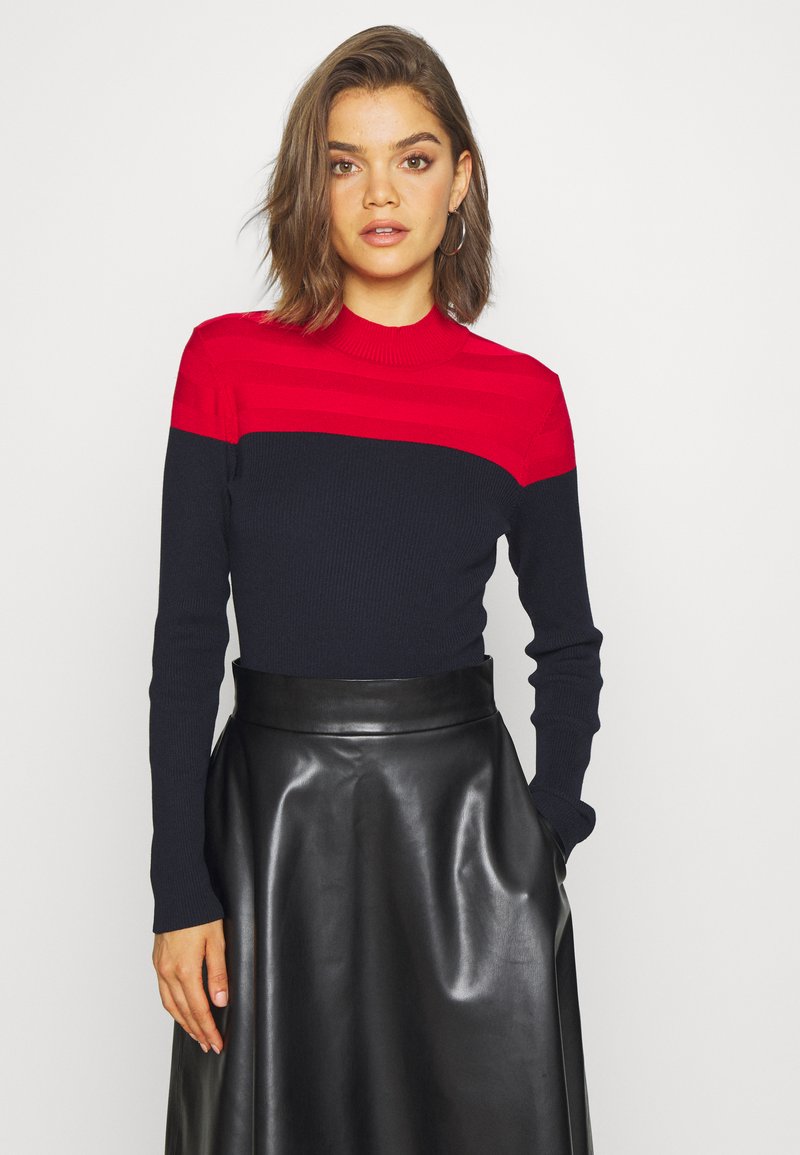 Un pull côtelé rouge et marine avec de longues manches et un col haut, associé à une jupe en cuir noir brillant. Texture lisse et design ajusté.