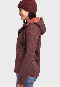 Rote und burgunderfarbene gemusterte Fleecejacke mit einer pinken Kapuze, verstellbaren Zugkordeln und einer seitlichen Reißverschlusstasche. Eine gerippte Strickmütze rundet das Outfit ab.
