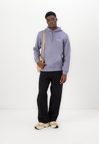 Hoodie en polaire grise avec une poche kangourou et un logo blanc. Associé à un pantalon noir à jambes larges et des baskets beiges. Porte un petit sac marron clair.