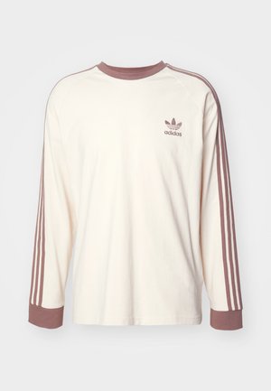 Camicia a maniche lunghe di colore crema chiaro con maniche raglan, polsini marroni scuri e tre strisce marroni scure su ciascun lato. Presenta un logo ricamato sul petto.