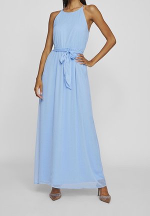 Kvinde iført ærmeløs, lyseblå maxikjole med bælte bundet i taljen og glitrende spidse hæle, stående med den ene hånd på hoften.