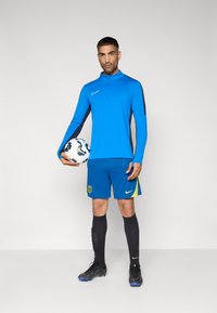 Μπλε αθλητικό μακρυμάνικο τοπ με φερμουάρ τριών τετάρτων και μαύρες πλαϊνές λωρίδες, συνδυασμένο με μπλε σορτς με κίτρινη λεπτομέρεια. Μαύρα ποδοσφαιρικά παπουτσάκια.