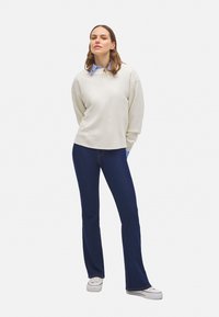 Sudadera oversized beige clara con escote redondo, combinada con jeans acampanados azul oscuro y zapatillas blancas, de pie frente a un fondo liso.